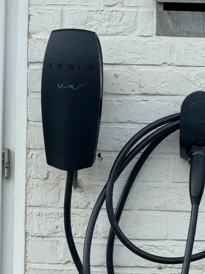 Zwarte Tesla Wall Connector aan een witte bakstenen muur, geplaatst door Laadnatie.