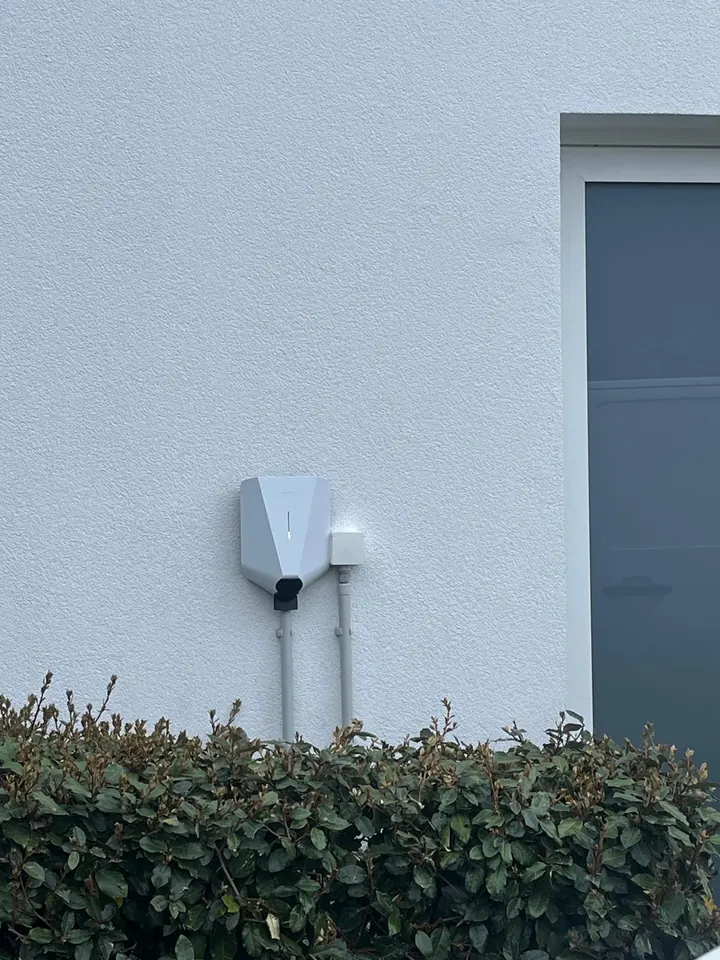 Witte Easee-lader aan een witte buitenmuur achter een haag, geplaatst door Laadnatie.